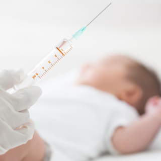 يد شخص تمسك حقنة وبداخلها سائل شفاف وطفل رضيع مستلقي على السرير