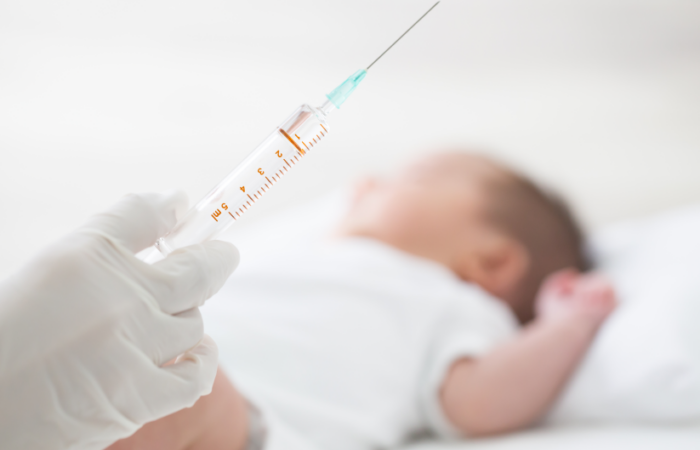 يد شخص تمسك حقنة وبداخلها سائل شفاف وطفل رضيع مستلقي على السرير