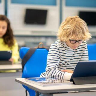 صبي شعره أشقر يجلس على كرسي أمام طاولة عليها أيباد ضمن قاعة صفية