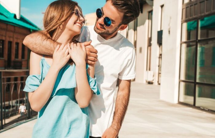 رجل يرتدي نظارة شمسية ويضع يده خول عنق امرأة مبتسمة تنظر لوجه
