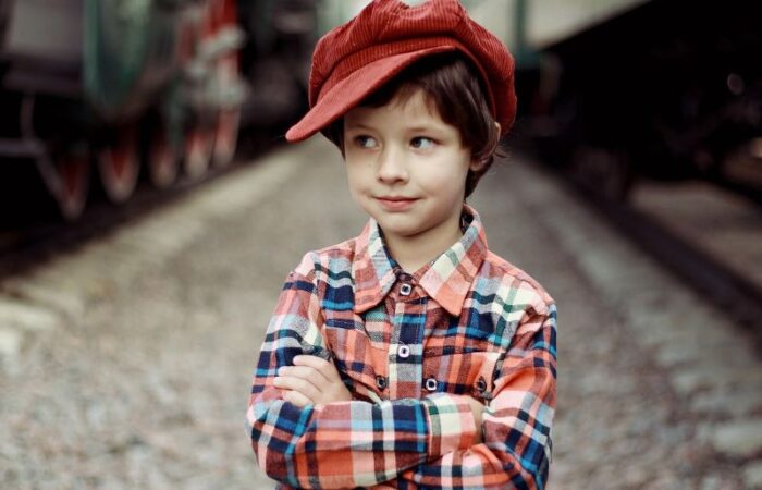 طفل يرتدي قميص متعدد الألوان ويضع قبعة حمراء على رأسه ويقف متشابك الأيدي بين قطارين بخاريين