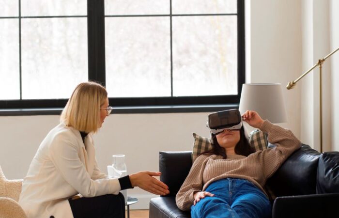 امرأة تجلس على الكرسي وتنظر لامرأة مستلقية على الأريكة وترتدي نظارة الواقع الافتراضي