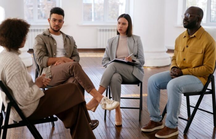 امرأة تجليس عل كرسي وتمسك قلم ودفتر بيدها ويجلس حولها رجلان وامرأة