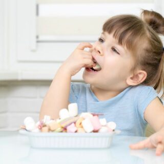 بنت تجلس على كرسي وبيدها قطعة سكاكر تضعها في فمها وأمامها صحن ممتلئ بالسكاكر