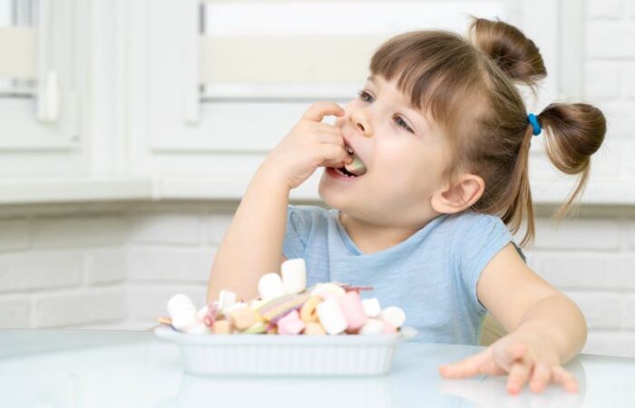 بنت تجلس على كرسي وبيدها قطعة سكاكر تضعها في فمها وأمامها صحن ممتلئ بالسكاكر