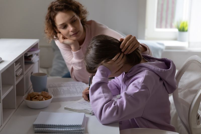 فتاة تجلس على كرسي أمام طاولة عليها كتب ودفاتر وتضع يدها على رأسها وبجانبها امرأة