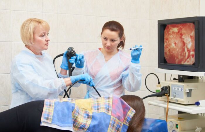 طبيبة تقوم بإدخال جهاز التنظير في فم مريضة مُستلقية على السرير وتقوم ممرضة بمساعدتها وتوجد أمام الطبيبة شاشة تعرض شكل الأعضاء من الداخل