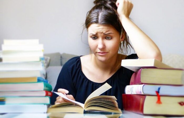 شابة تضع يدها على رأسها ويبدو عليها التوتر تجلس على كرسي وأمامها طاولة عليها كتب كثيرة