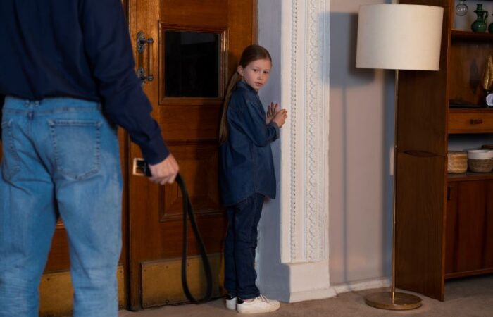 رجل بالغ يمسك بيده حزام البنطال وتقف أمامه بنت تقف خائفة بجانب الباب