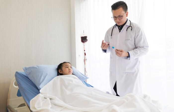 فتاة صغيرة ممددة على السرير في المستشفى وبجانبها عمود بأعلاه كيس دم وطبيب يمسك إبرة بداخلها سائل أحمر