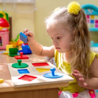 فتاة صغيرة من ذوي الاحتياجات الخاصة تقوم باللعب بقطع هندسية ملونة موجودة أمامها على الطاولة