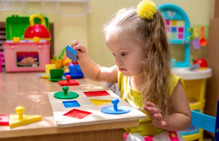 فتاة صغيرة من ذوي الاحتياجات الخاصة تقوم باللعب بقطع هندسية ملونة موجودة أمامها على الطاولة