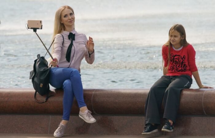 امرأة شابة تجلس على رصيف الشاطئ بجانب البحر وتمسك عصا سيلفي لتصوير نفسها وبجانبها فتاة حزينة