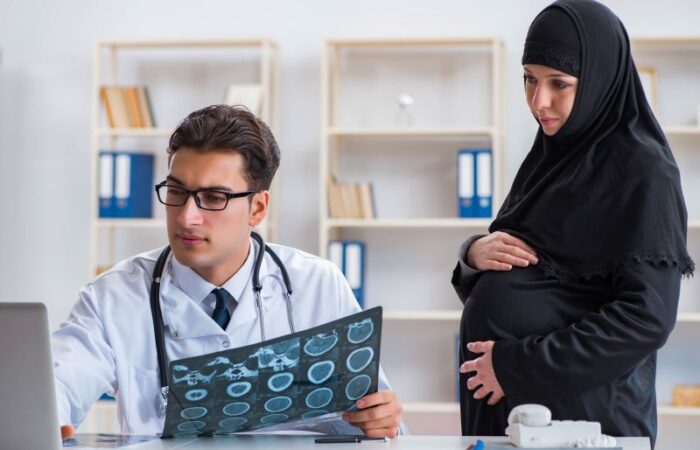 امرأة حامل تضع يدها على بطنها وبجانبها طبيب يجلس على كرسي ويمسك بيده صورة أشعة