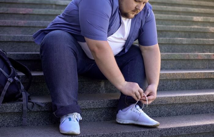 رجل بالغ بدين يجلس على الدرج ويحاول ربط شريط حذائه