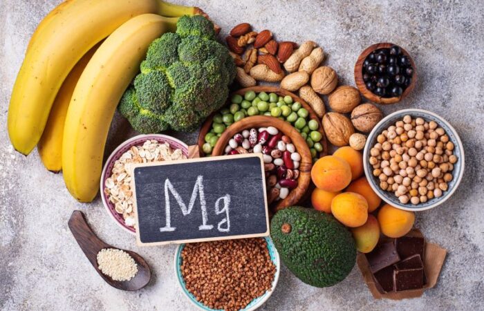 مجموعة من المأكولات: الموز، الأفوكادو، البروكلي، قطع من الشوكولا، وبقوليات كحبوب البازلاء والفاصولياء الحمراء والبيضاء ويتوسطها لوح أسود صغير مكتوب عليه بالإنكليزية Mg