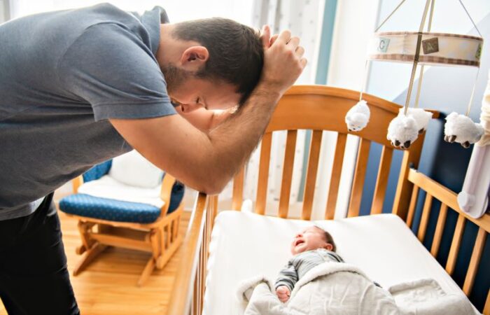 طفل رضيع مستلقي في سريره ورجل بالغ حزين يقف بجانب السرير ويضع يديه على رأسه