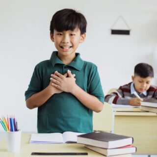 صبي يقف أمام طاولة في الصف ويضع يديه مضمومتين على صدره