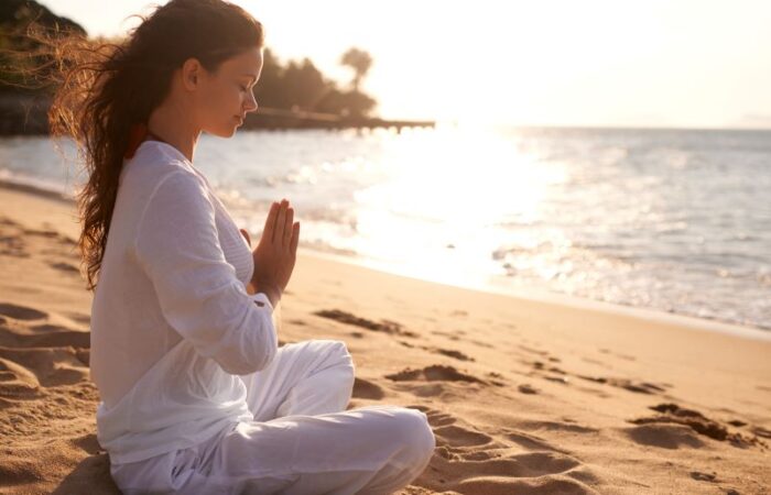 امرأة شابة تجلس على الرمل أمام شاطئ البحر مغمضة العينين وتضع يديها متطابقة أمام صدرها