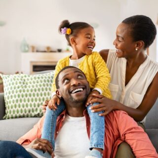 عائلة مؤلفة من ثلاثة أشخاص هم الأب والأم وفتاة يجلسون في غرفة المعيشة وتظهر الفتاة فرحة وهي تجلس على كتفي والدها