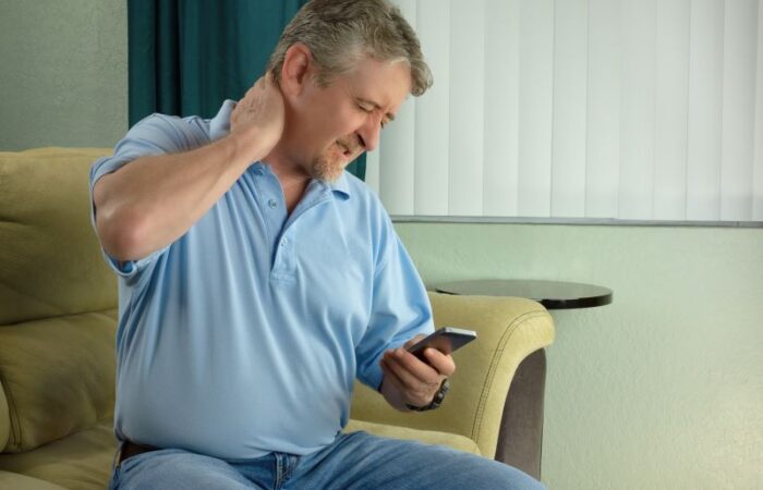 رجل بالغ يجلس على الأريكة يحمل بيده الهاتف المحمول ويده الأخرى يضعها على رقبته ويشعر بالألم