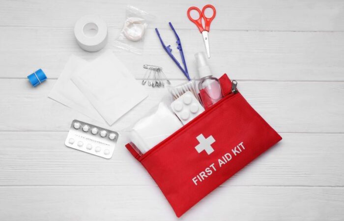حقيبة صغيرة حمراء اللون مرسوم عليها إشارة + وبجانبها يوجد مقص وضماد طبي وظروف أدوية