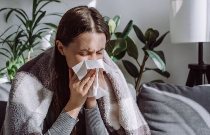 امرأة شابة تجلس على الأريكة وتضع منديل ورقي على أنفها وهي مغمضة العينين